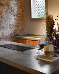 Cucina su misura in cemento e legno con isola e piano cottura a induzione, davanti a un muro in sasso recuperato