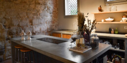 Cucina su misura in cemento e legno con isola e piano cottura a induzione, davanti a un muro in sasso recuperato